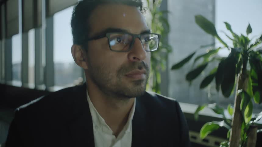 A man wearing glasses and a business jacket and dress shirt sits beside a wall windows and stares forward deep in thought on a bright day