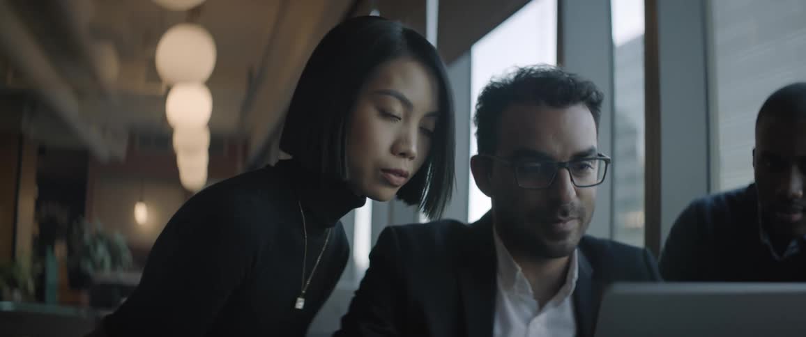 A woman is sitting next to a man wearing glasses who is looking at a laptop and talking to her
