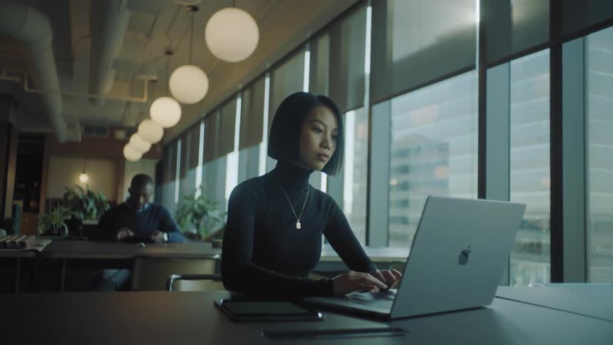 A woman sitting is using a laptop on a table and there are large glass windows behind her