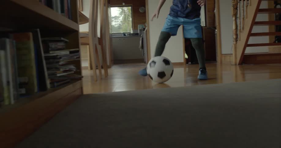 A young boy drops and kicks a soccer ball across a wooden floor indoors.