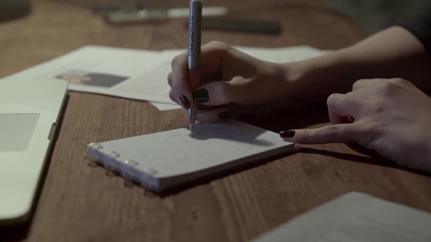 A woman writes in a small spiral notebook while working at a wooden desk.