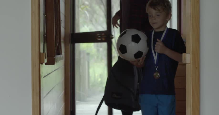 A boy carrying a soccer ball enters a house followed by his father