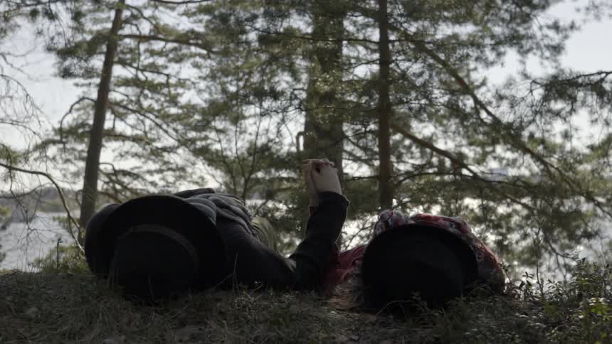 A man and woman lay lakeside in a forested area while holding hands