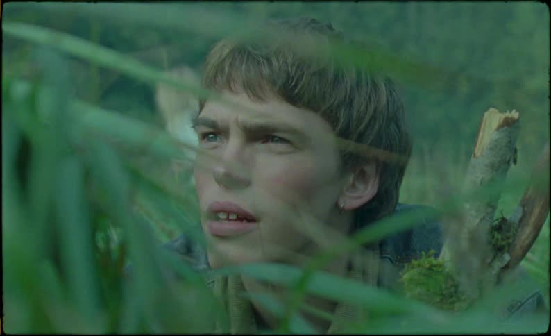 Two people in the forest with the boy squatting while holding a stick