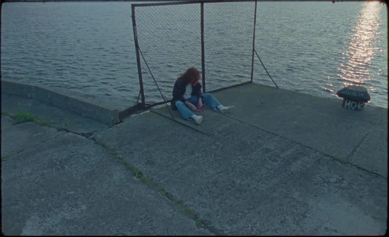 Person sits on concrete leaning against fence fiddling with things in their pockets