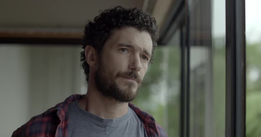 A man with a beard looks thoughtfully out of a large window while indoors.