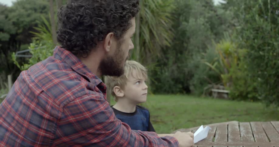 A man makes a white paper airplane for his young son sitting outdoors together.