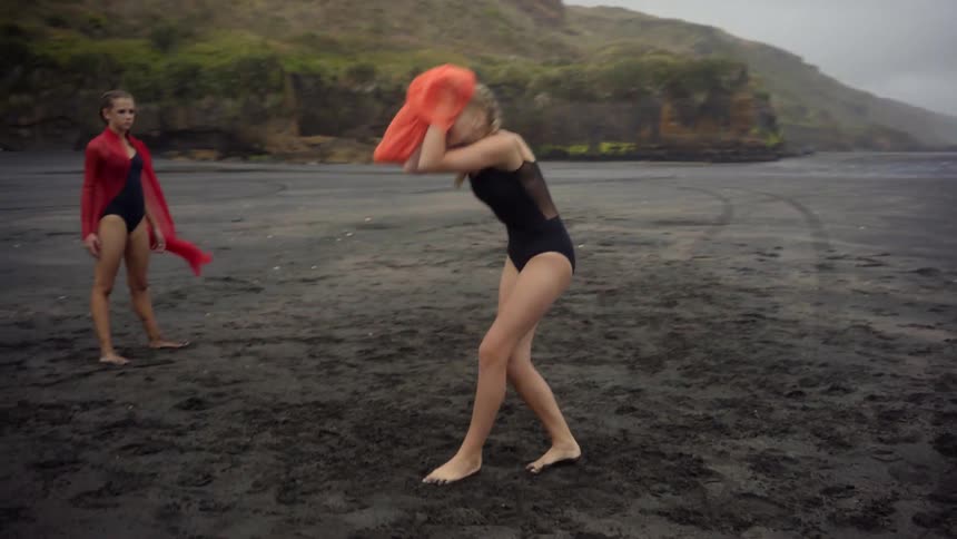A blonde girl holding an orange cloth dances on the beach while her friends watch
