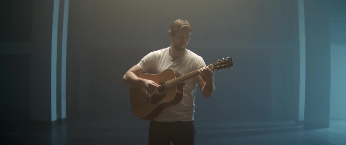 Young man strumming a wooden acoustic guitar while standing alone in a professional studio