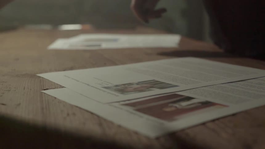 A creative professional organizes several project drafts and photos across a large wooden table.