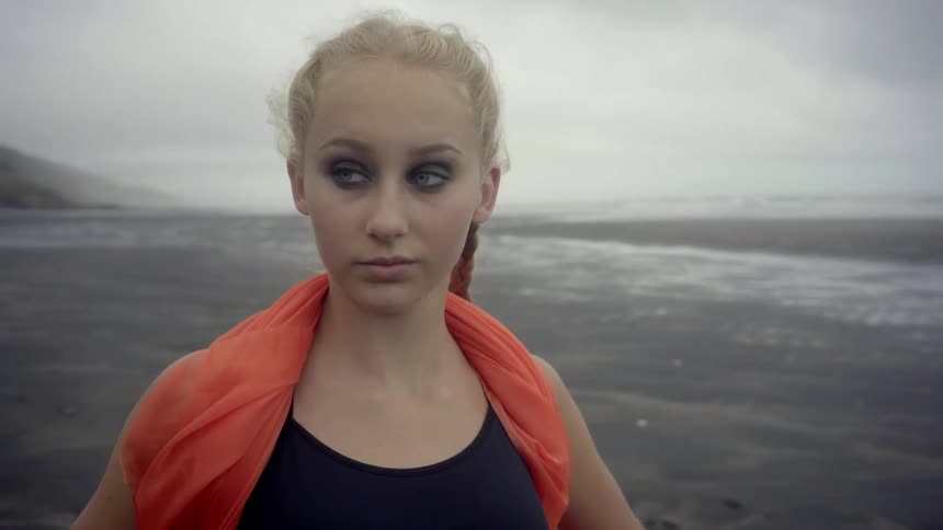A melancholic girl wearing an orange scarf gathers her thoughts by the powerful ocean.