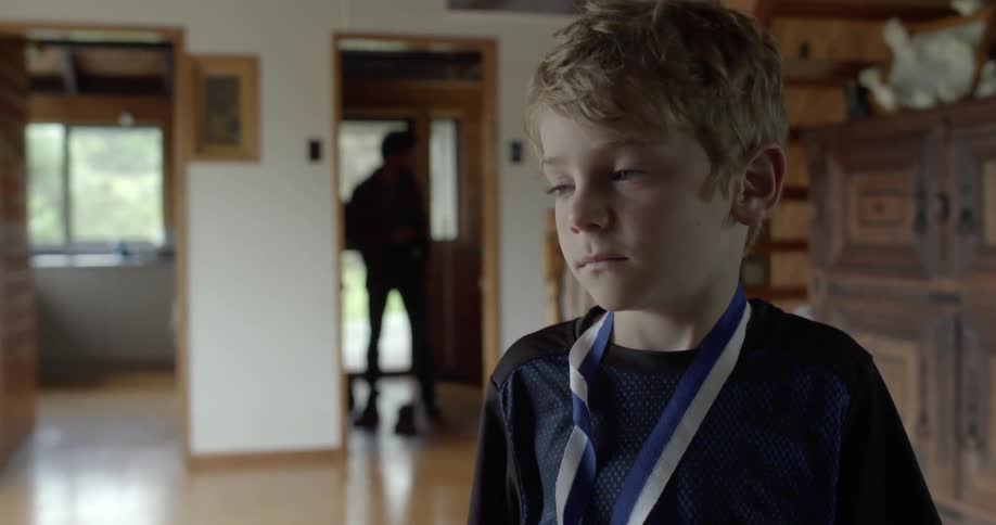 A young boy stands motionless as a man searches in the room behind