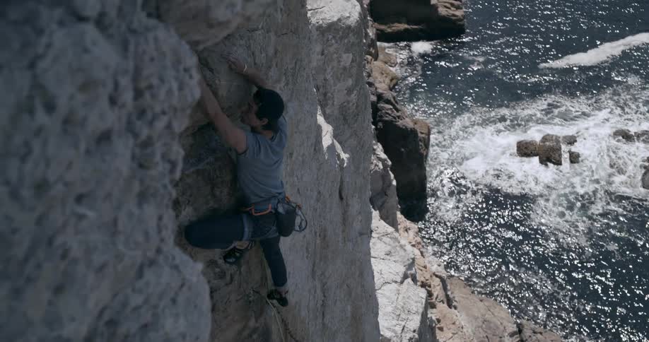 Male rock climber scaling a coastal cliff rock with sunlight reflecting from the water in the background