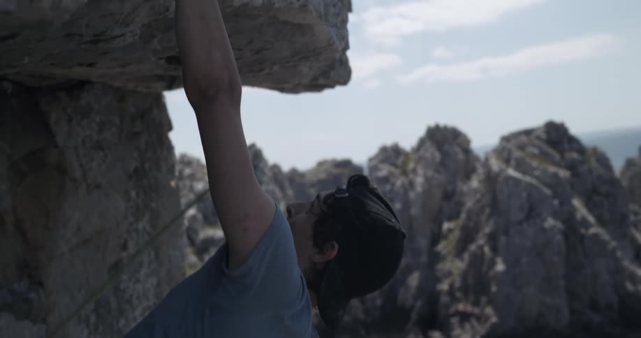 Young adult male hangs from and attaches a hook to a rock on a coastal cliff above water during the day