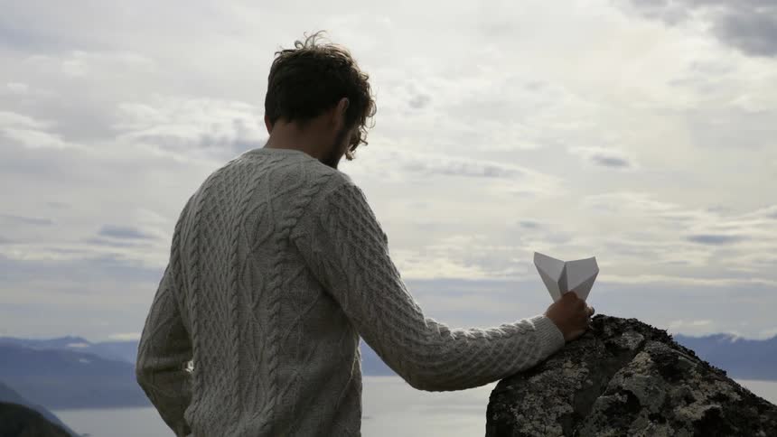 A nostalgic traveler prepares to launch a paper airplane across a scenic mountain valley.