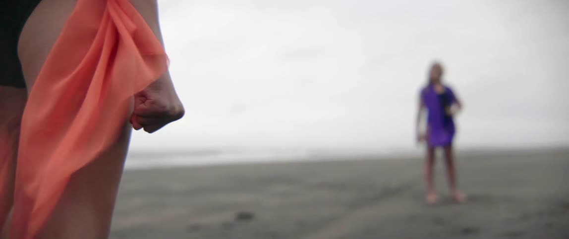A girl wearing a sheer orange cloth clenches her fist on the beach