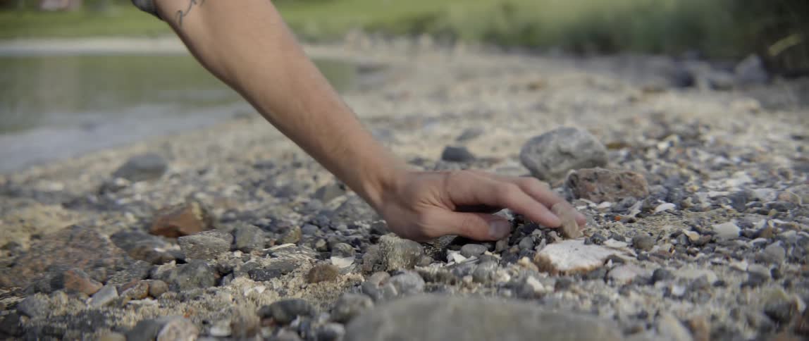 A man uses his hand to brush rocks and sand on the shoreline