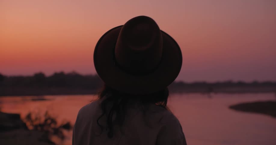 Woman wearing a hat looks at a river at sunset