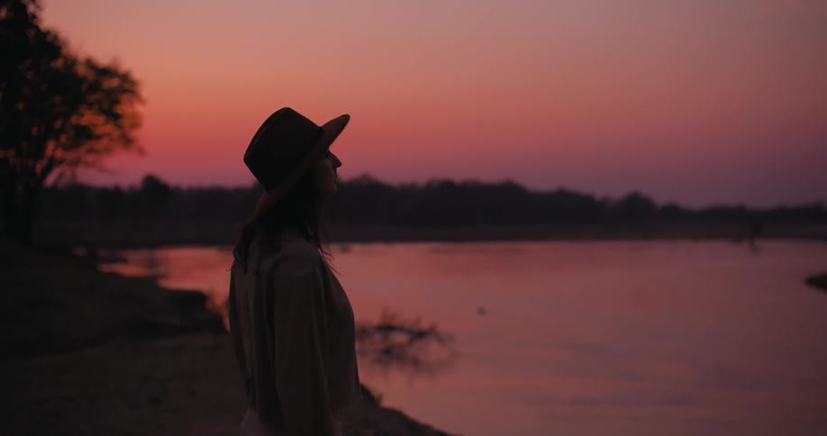 Woman wearing a hat stands on the coast of a lake at sunset