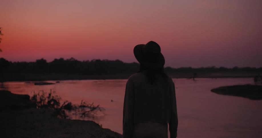 Woman wearing a hat stands near a lake at sunset