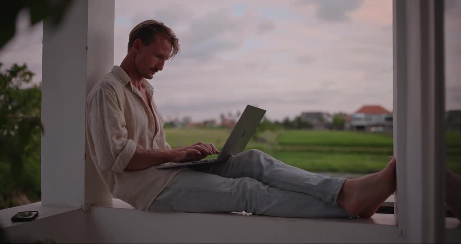 A man working on his laptop on the porch of a house in a rural town