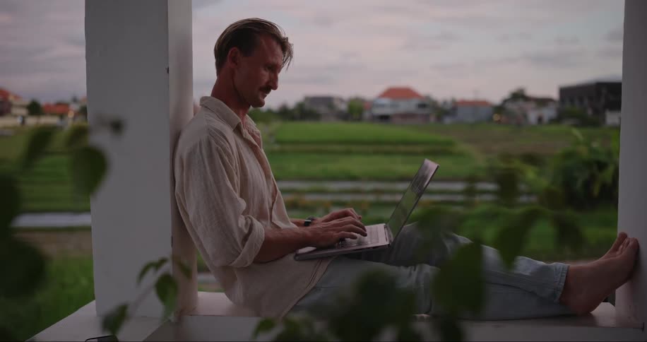 A man sits outside on his porch typing on a laptop computer
