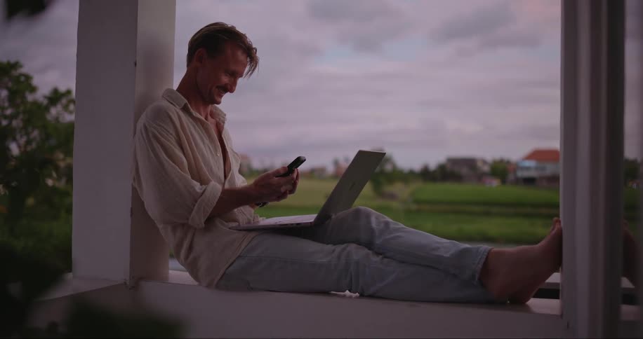 A man is smiling talking on his phone and working on his laptop while sitting on the balcony