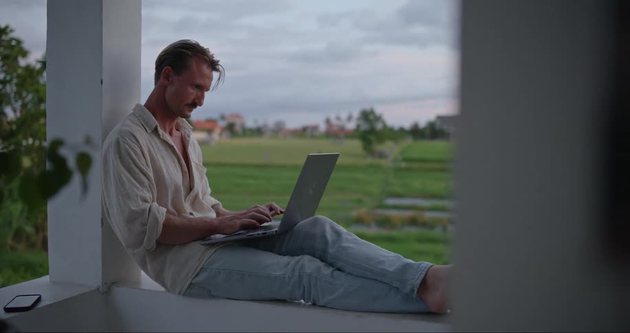 A man sitting on the balcony thinking and typing on his laptop