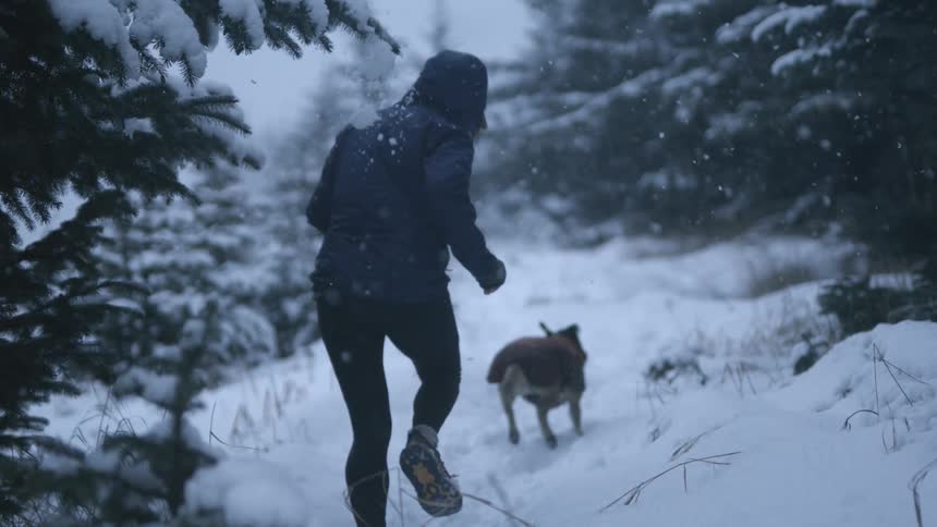 A woman running through a snow covered pine forest behind a pet dog in slow motion as snow falls around them