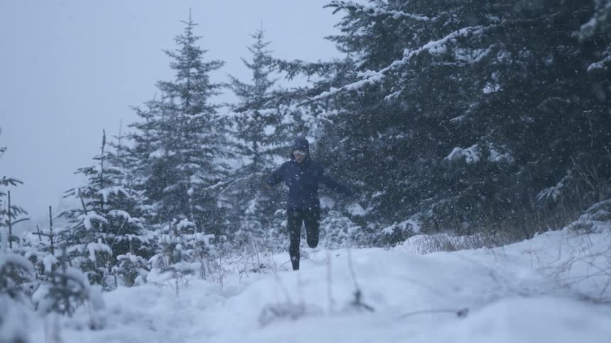 Woman running in slow motion through the snow with snow covered pine trees behind her as snow falls in a white haze all around