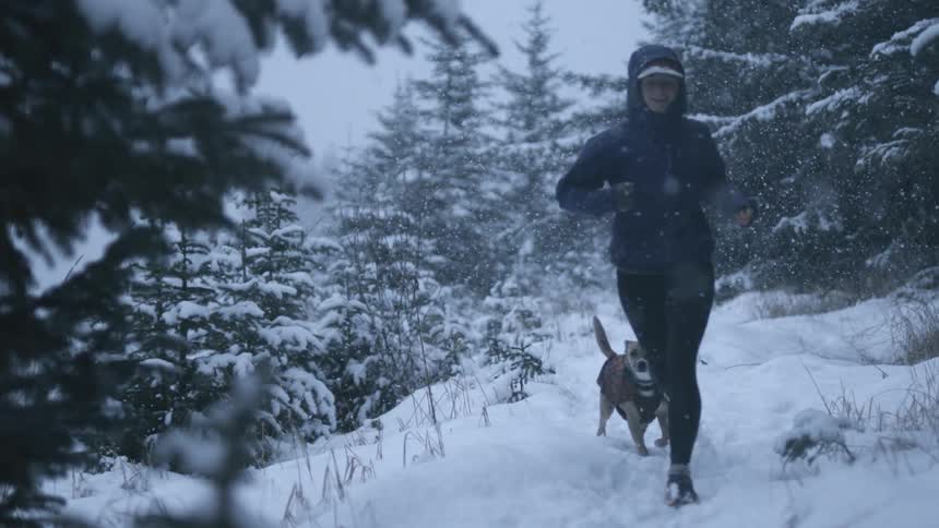 A young woman runs along a forested trail with her dog on a snowy winter day