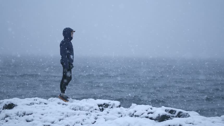 Woman standing on a snow covered rocky shoreline beside a wavy sea as snow falls with a clouded hazy sky in the background
