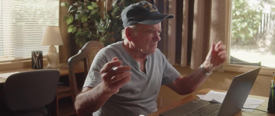 An elderly man is sitting at a table with a laptop computer and paperwork