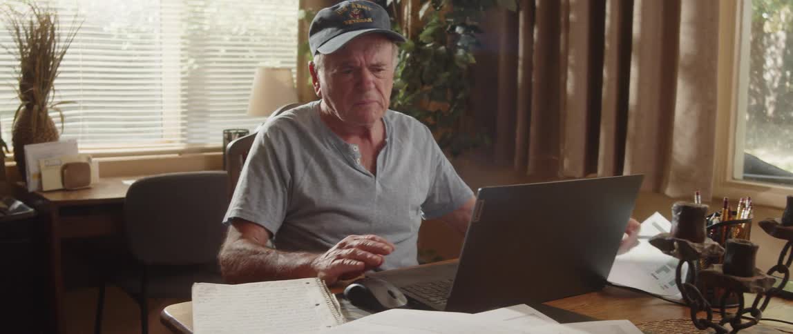 An elderly man is sitting at a table with a laptop computer and looking at paperwork