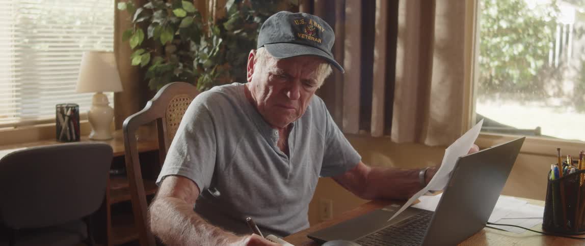 An elderly man is sitting at a table with a laptop computer doing paperwork