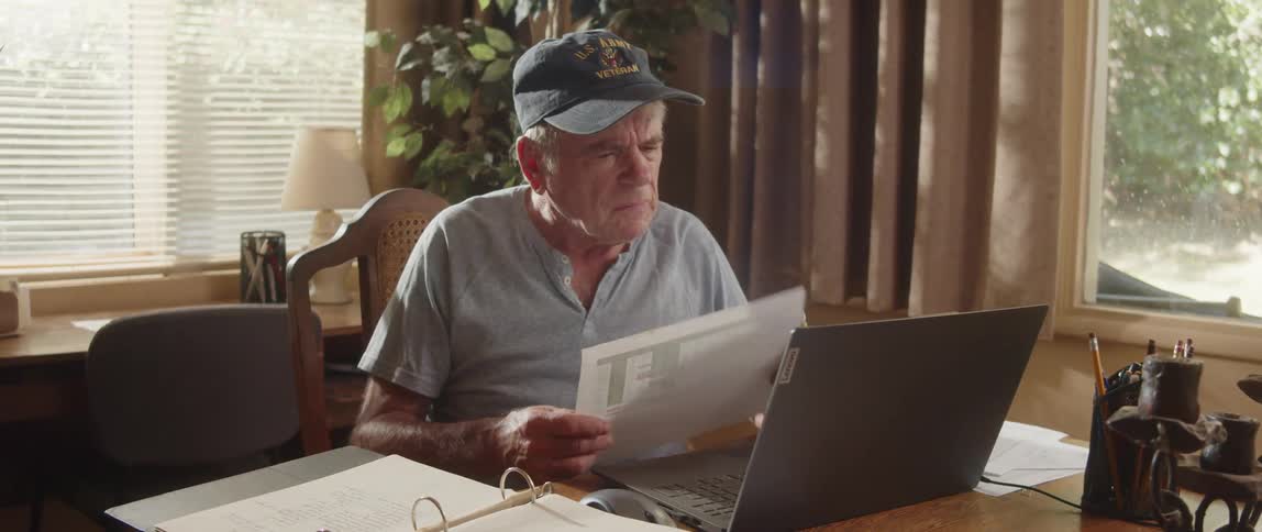 An elderly man is sitting at a table with a laptop computer and looking at paperwork