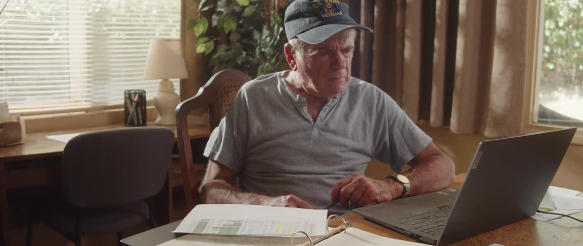 An elderly man is sitting at a table with a laptop computer and doing paperwork