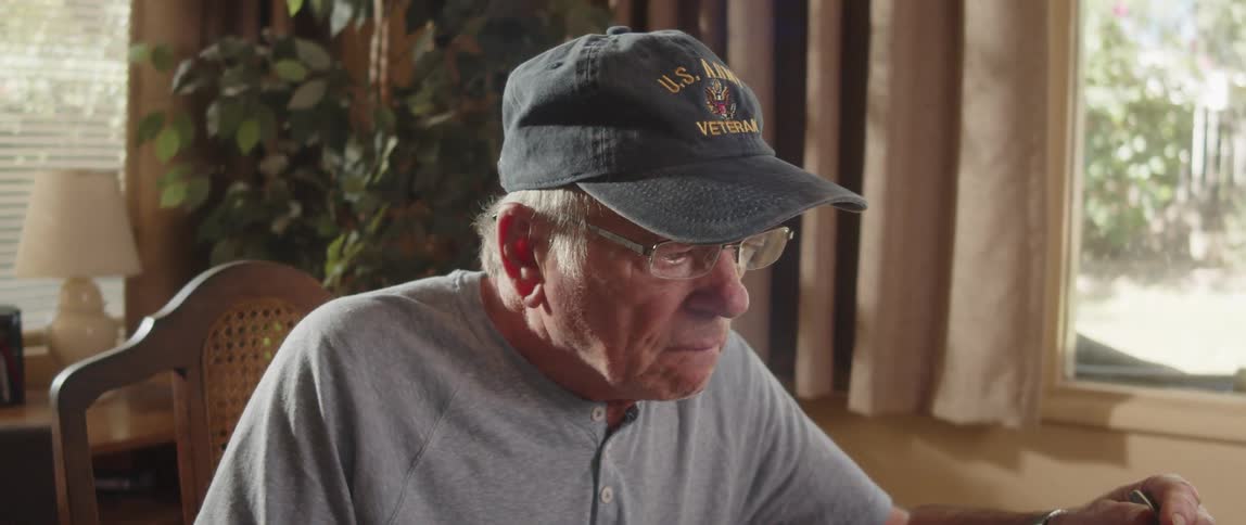An elderly man wearing a cap and reading glasses is looking at something in front of him inside a room