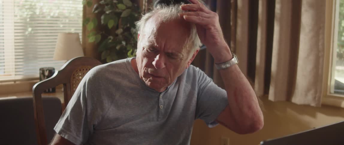 An elderly man is sitting at a table with a laptop computer and looking at paperwork and he takes off his cap as he thinks