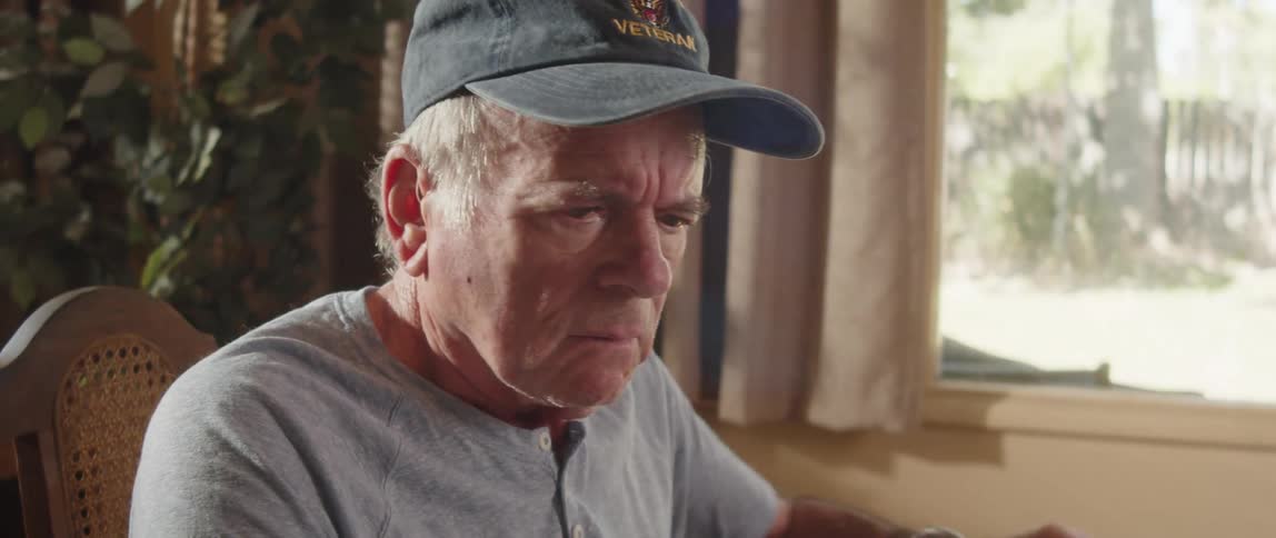An elderly man in a baseball cap reviews materials and pauses to contemplate