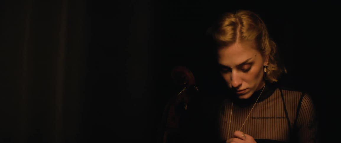 A woman stands with her cello in the darkness behind a curtain and nervously paces
