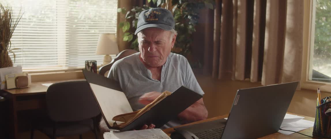An elderly man is sitting at a table with a laptop computer going through paperwork in a notebook