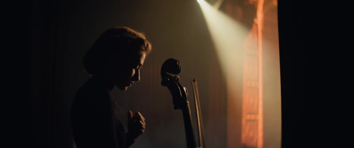 A nervous woman holds her necklace and cello right before walking into the stage spotlight