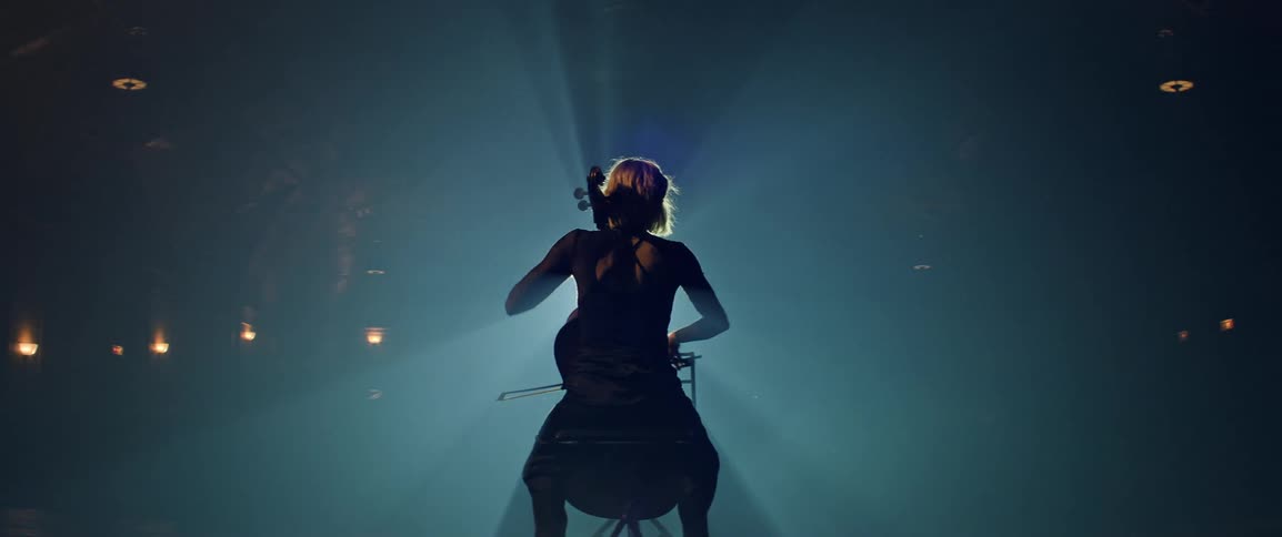 Young blonde female soloist plays the cello in the flare of a large stage spotlight