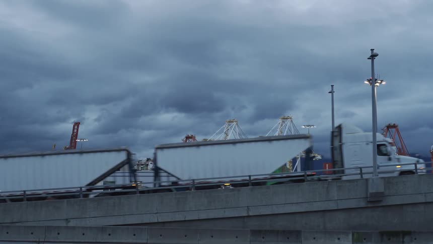 Semi truck hauling multiple containers crosses bridge near manufacturing plant on an overcast day