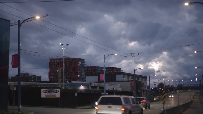 Vehicles move along highway past apartment buildings under an overcast sky at night