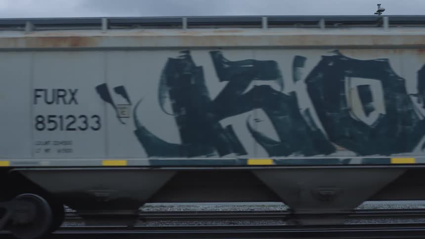 Graffiti covered train cars travel in the opposite direction of a semi truck with a factory in the background
