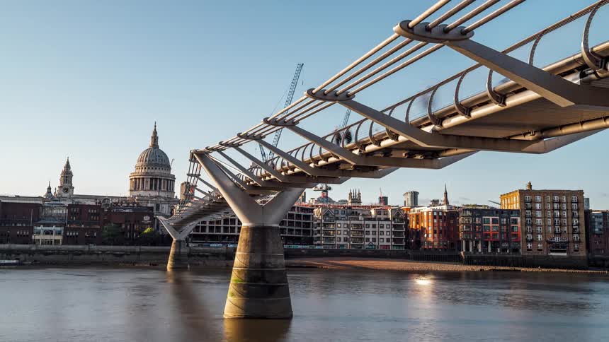 Timelaps people crossing the millennium bridge over the river thames in london with the city waterfront in the background at sunset