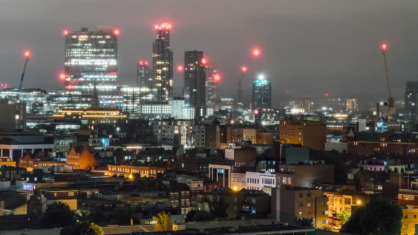 A city with several buildings and houses with lights on and the clouds in the sky moving quickly