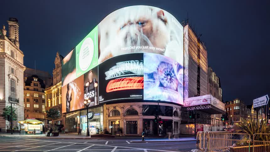Pedestrian and vehicular traffic moves through intersection passing building with large outdoor digital screens